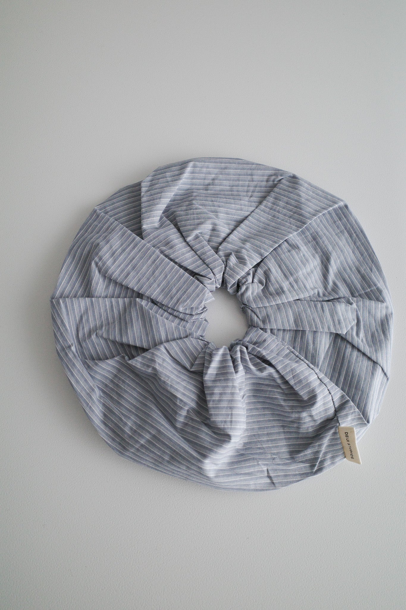 A large, round Scrunchie made of light blue and white pinstriped fabric with a small tag attached is laid flat on a light gray surface.