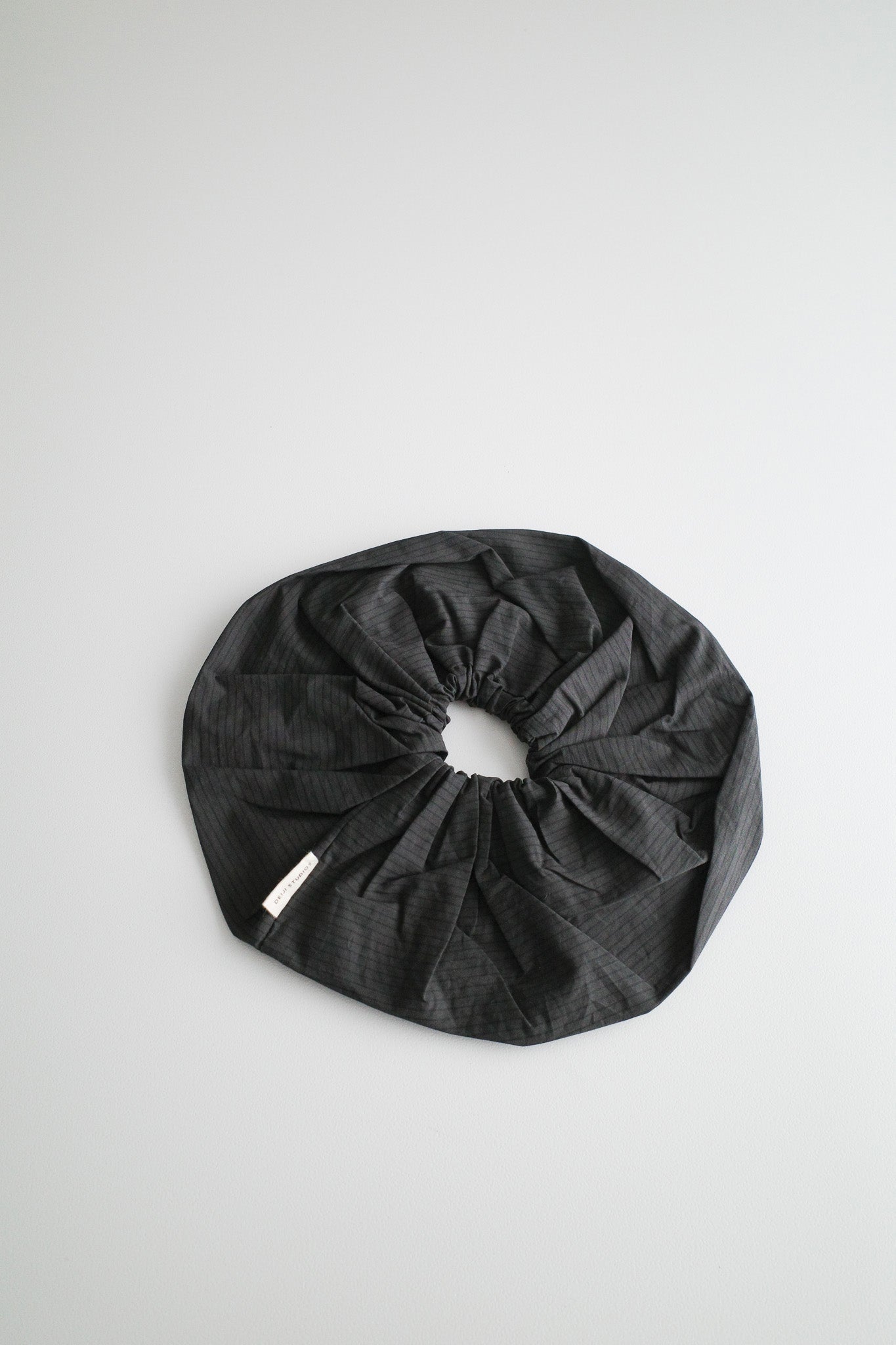 The Scrunchie - Black Stripe, an oversized dark gray hair accessory with subtle stripes, lies flat on a light gray surface. A small white tag is attached to one side, highlighting Deiji Studios’ signature style.