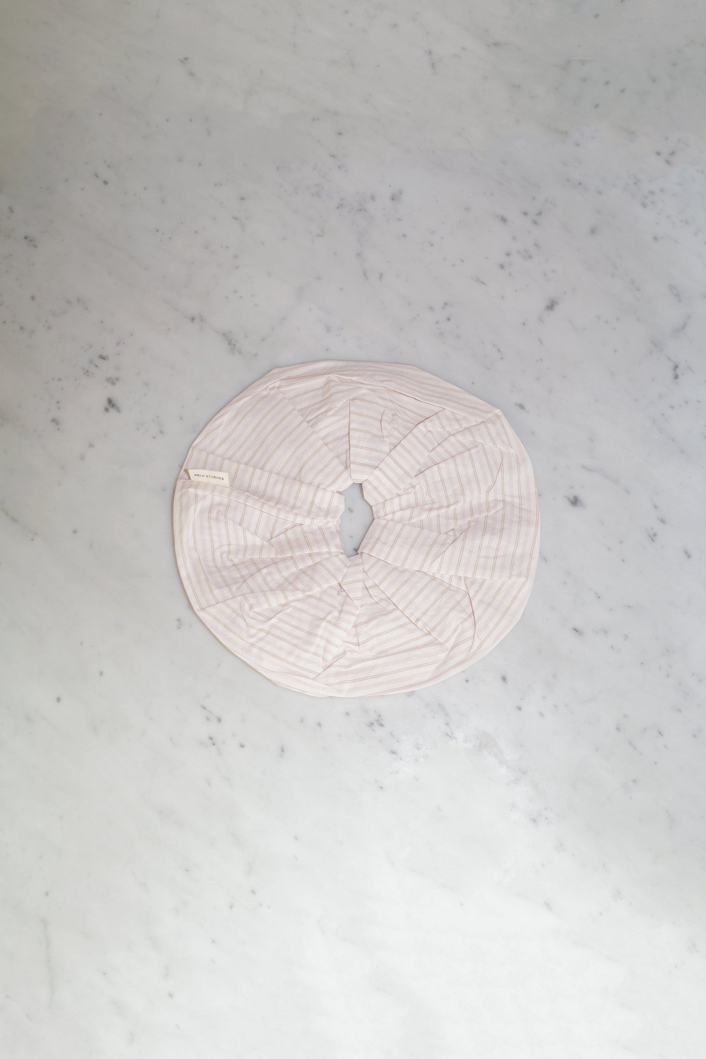 A light pink, striped fabric Scrunchie with a circular shape and gathered elastic center, displayed on a white marble surface.