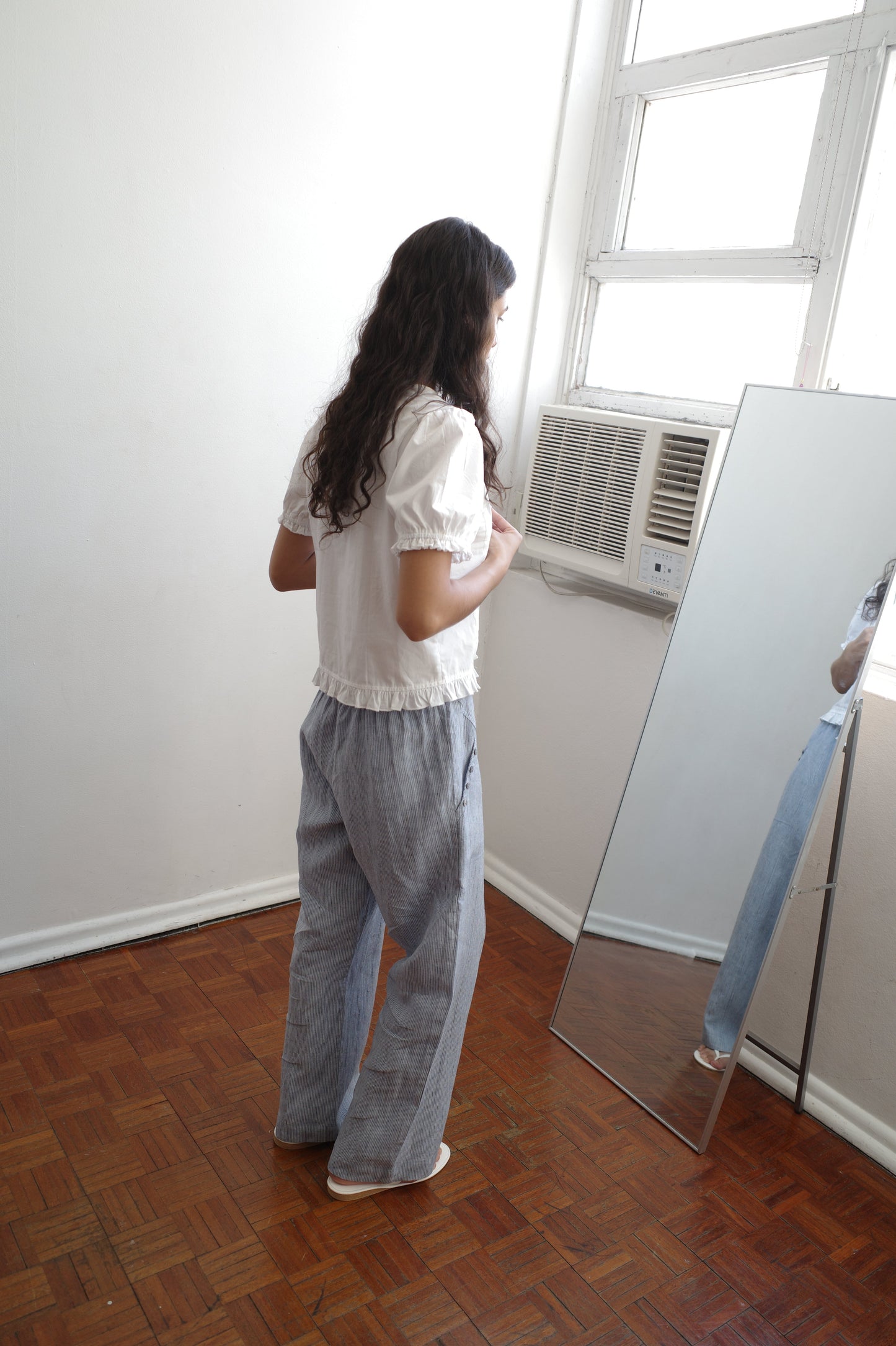 A person with long dark hair, wearing the Pintuck Top - White from Deiji Studios and loose striped pants, stands on wooden floors gazing at their reflection in a tall mirror near a window and air conditioner.