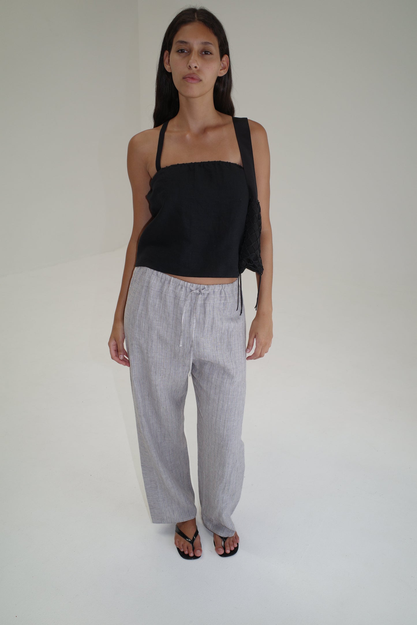 A woman with long dark hair wears a black sleeveless top, Double Waist Linen Pant in Haze Blue, and black sandals, standing in a minimalist white room and looking at the camera with a neutral expression.