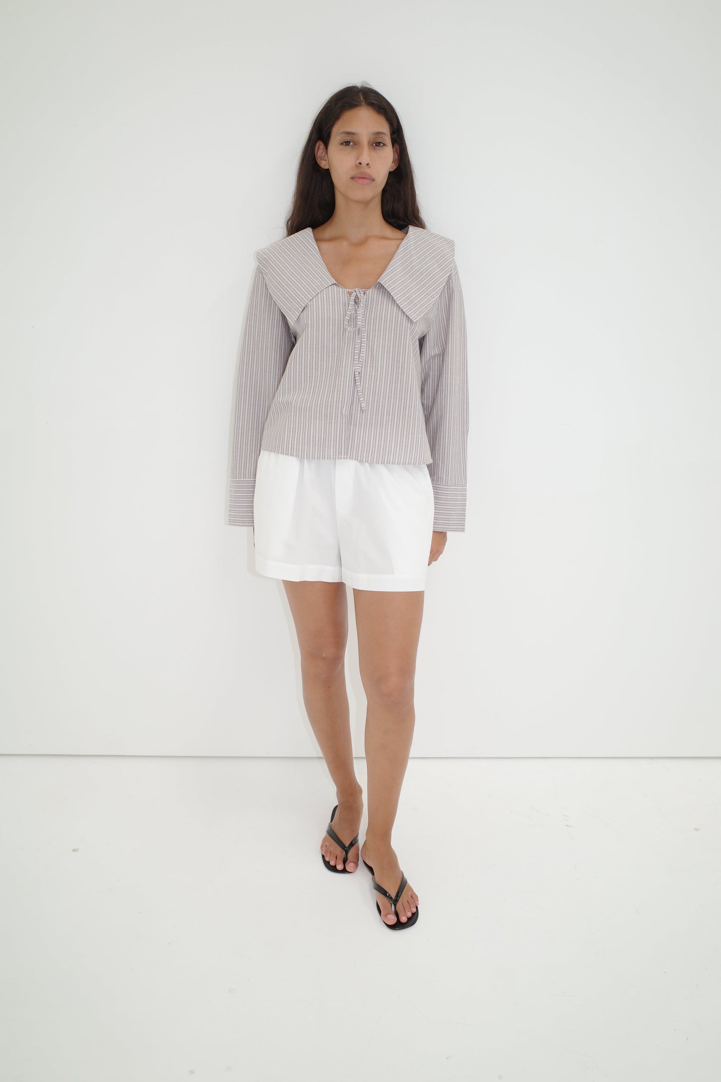 A woman with long dark hair and a neutral expression stands against a white wall, wearing the Collared Tie Shirt - Seas Stripe—an oversized shirt with bold stripes and a large collar—paired with white shorts and black flip-flops.