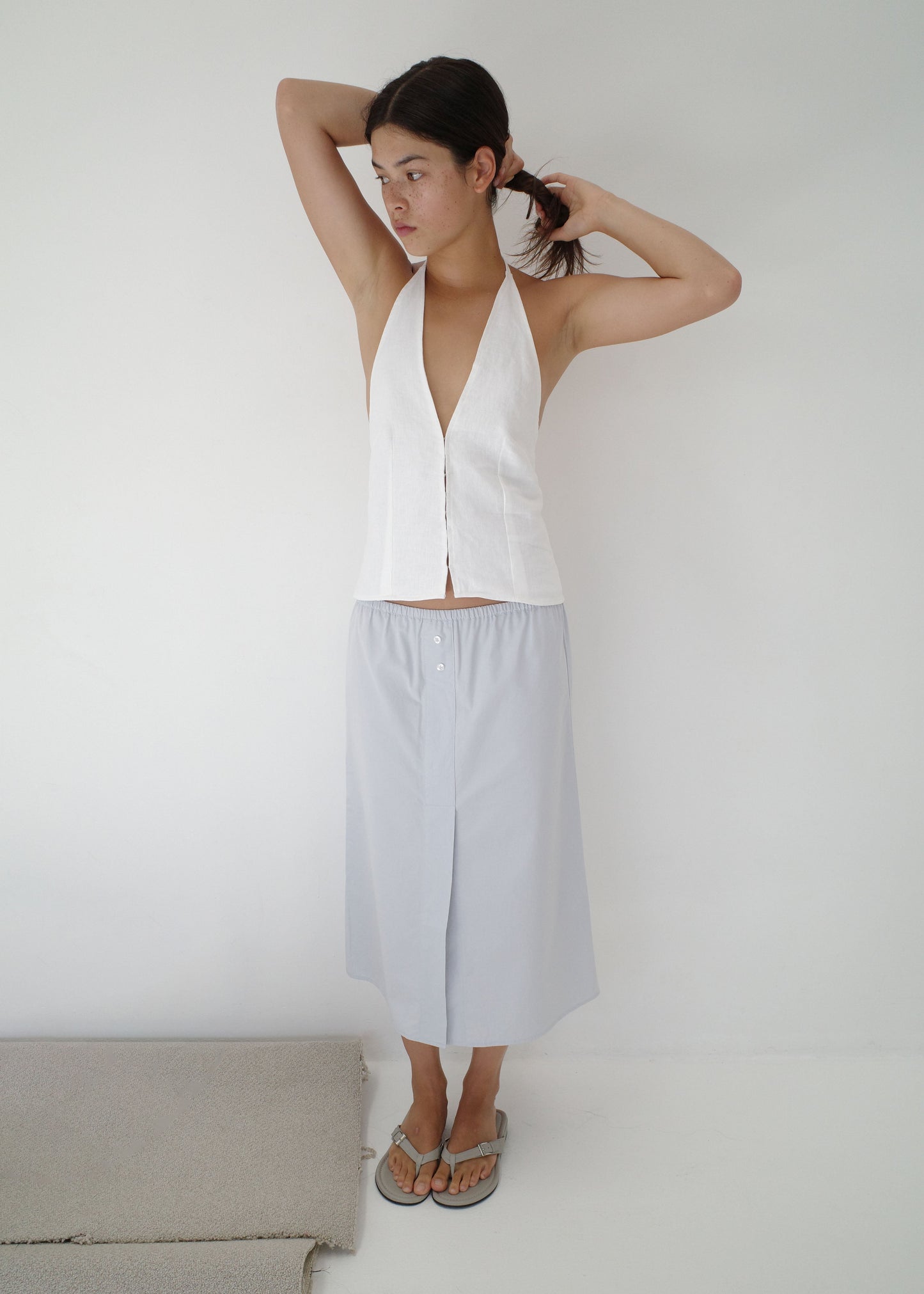 A person with dark hair stands against a white wall, adjusting their hair while wearing a sleeveless white halter top, flip-flops, and the Overlap Skirt in light gray that falls to the knee.