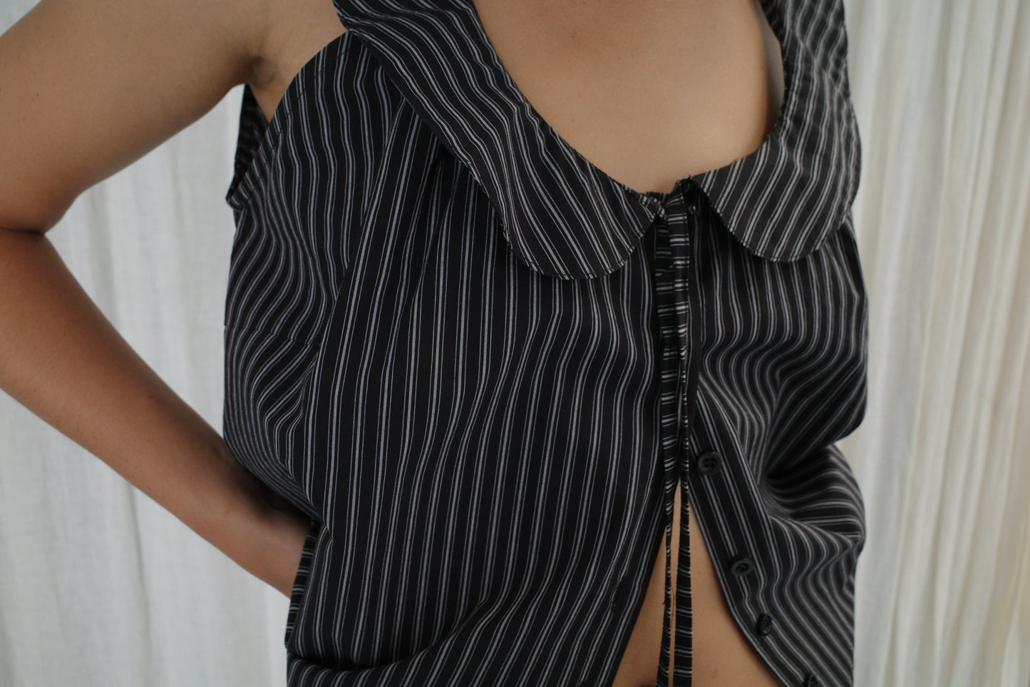 Wearing the Round Collar Top - Mono Stripe by Deiji Studios, which has black and white pinstripes, a large collar, and front ties, a person stands with one hand on their hip against a light fabric background.