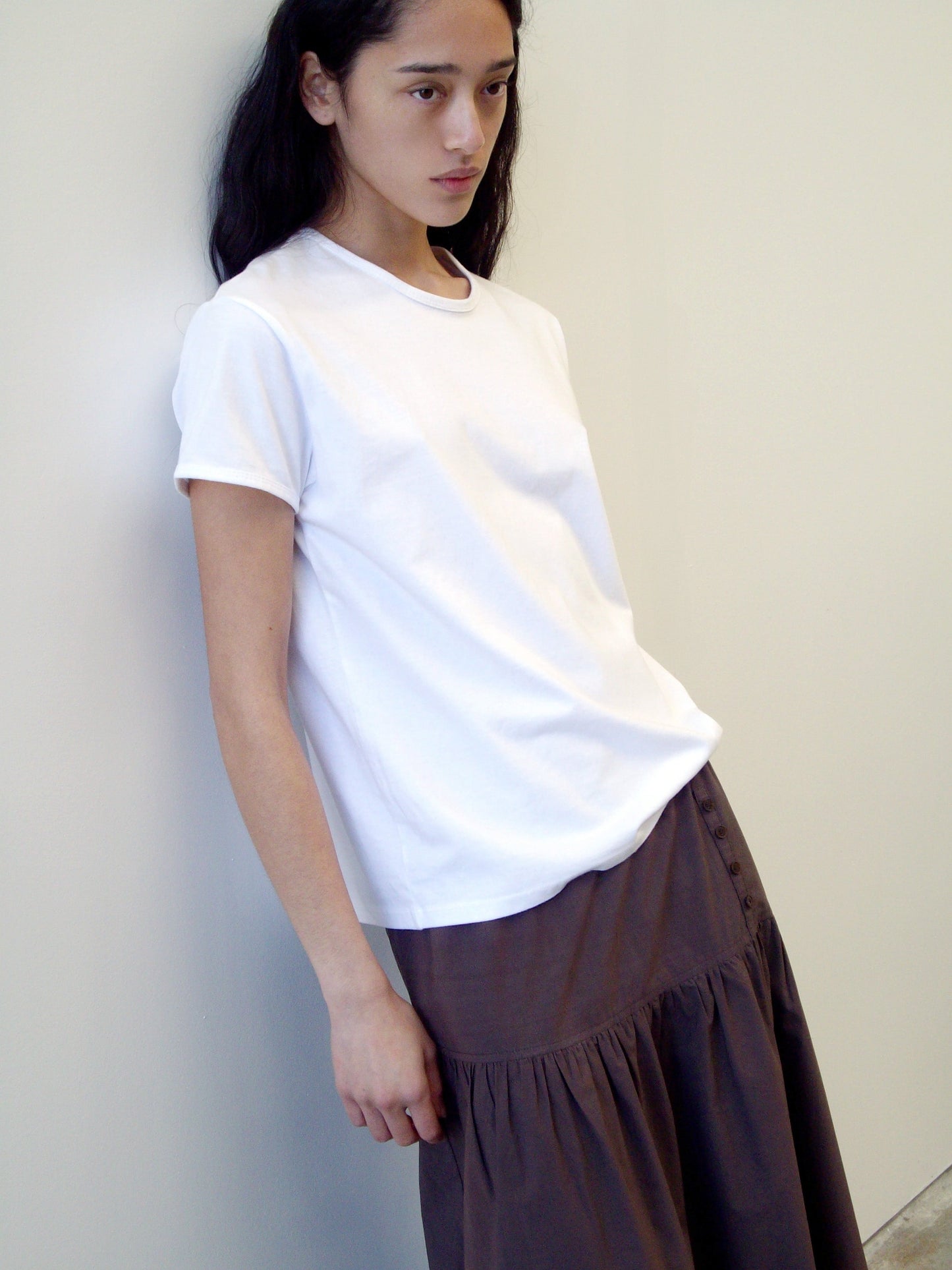 A person with long dark hair wears a plain white organic cotton t-shirt and the Button Skirt - Chocolate, leaning against a light-colored wall with a neutral expression.