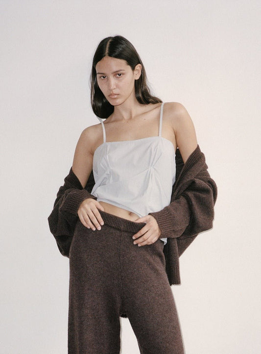 A person with long dark hair models the Pleat Top, a white camisole style, paired with a brown knit cardigan and pants, posing with one hand on their hip against a plain light background.