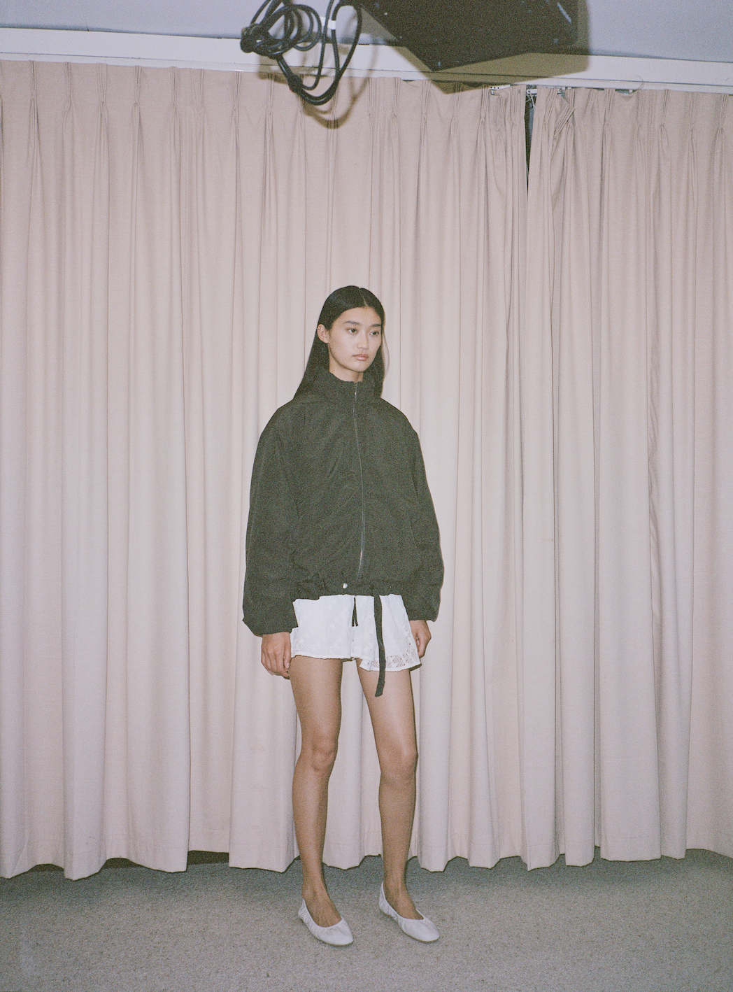 A woman with long dark hair stands indoors in front of beige curtains, wearing the 0 Press Stud Jacket in Dark Chocolate, paired with white shorts and white flats. A light fixture is partially visible at the top of the image.