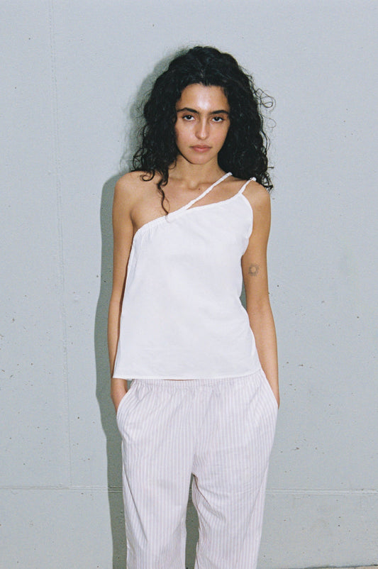 A person with long curly hair wears the Shoulder Top - White by Deiji Studios and loose, light-striped pants, standing against a plain light gray wall with hands in pockets.