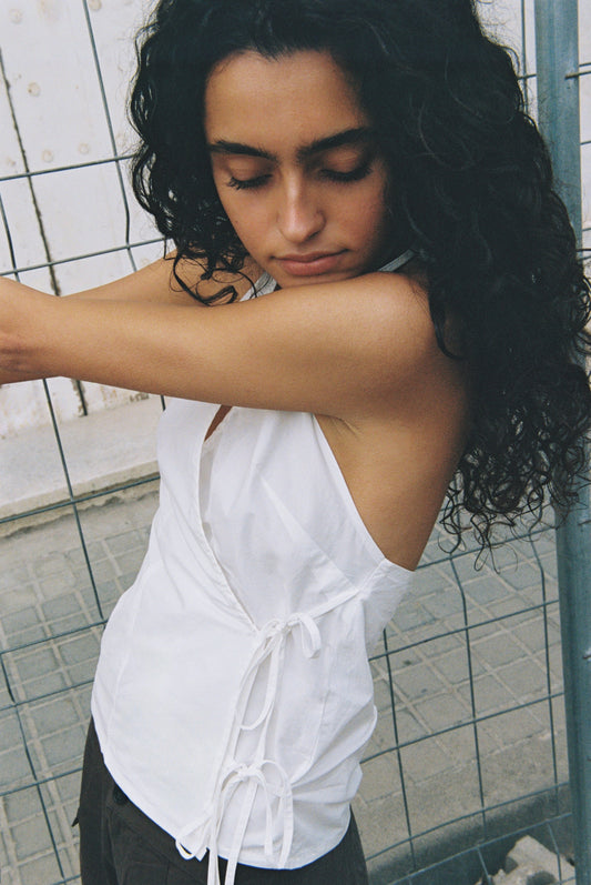 A woman with long, curly dark hair and closed eyes stands by a wire fence in a white Drawcord Halter and dark pants, one arm outstretched as she looks downward peacefully.