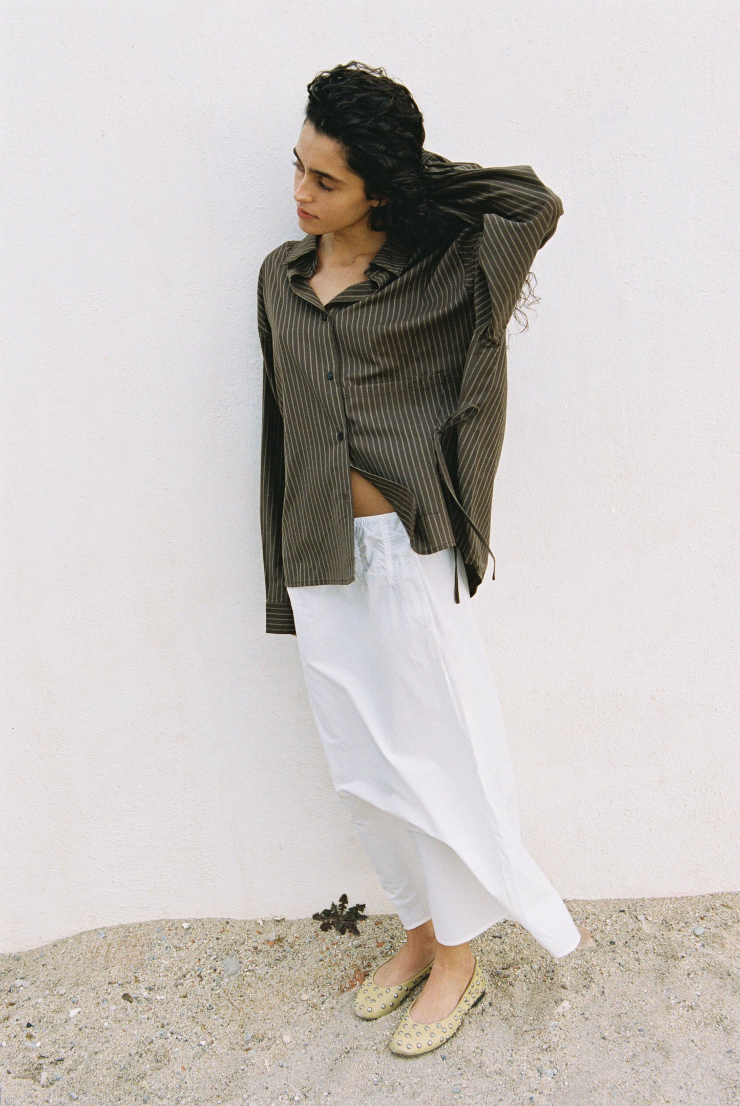A person with curly dark hair stands against a white wall, wearing the Pocket Wool Shirt with stripes and a long white skirt, paired with light-colored flats, looking down as they touch their hair.