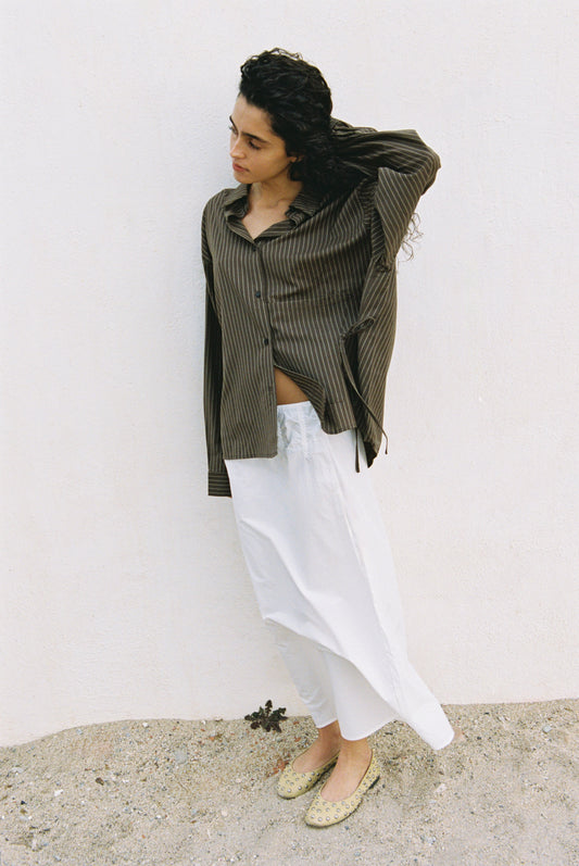 A person with curly dark hair stands against a white wall, wearing the Pocket Wool Shirt—a loose brown striped top. They pair it with a long white skirt and light flats, looking down as they brush their hair back with one hand.