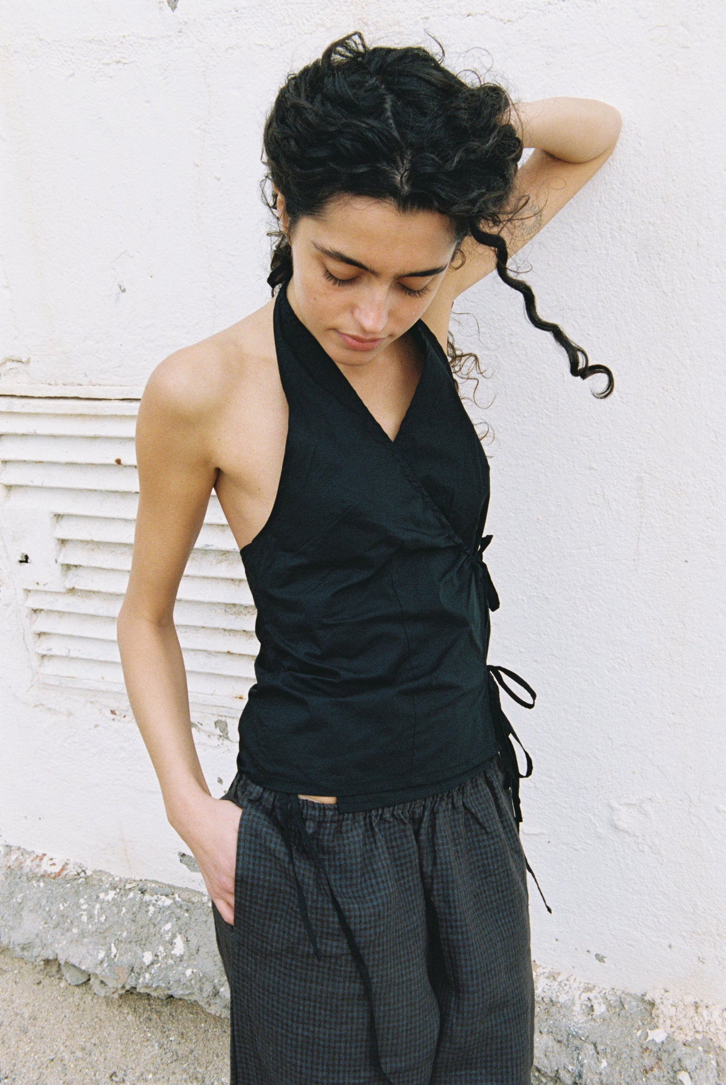 A woman with curly dark hair stands against a white wall, looking down with one arm raised and the other in her pocket. She wears the sleeveless black Drawcord Halter top paired with dark plaid pants.