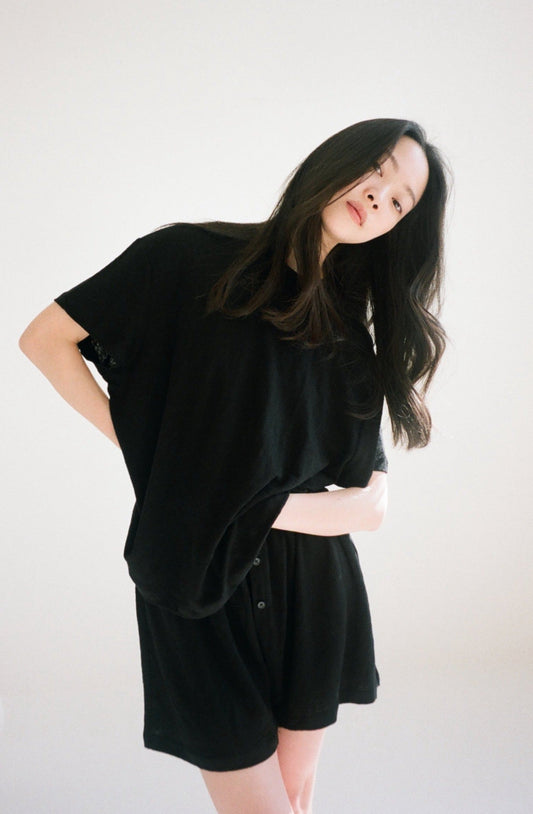 A woman with long dark hair models the Soft T-Shirt in black and matching shorts, standing against a plain light background and looking slightly downward at the camera.