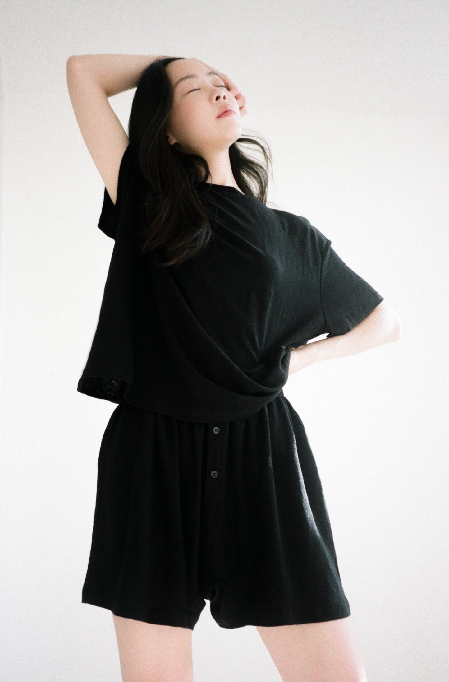 A woman poses confidently with one arm raised, eyes closed, wearing Soft Short and a loose black t-shirt against a bright, neutral background.