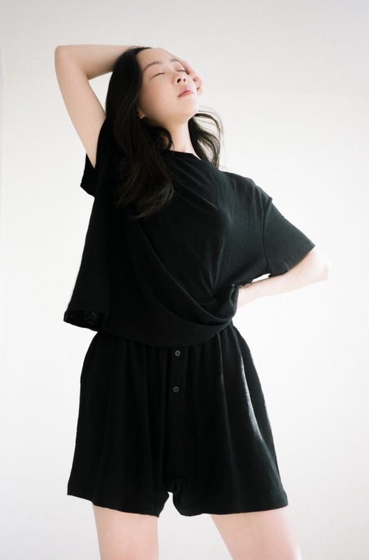A woman poses confidently with one arm raised, eyes closed, wearing Soft Short and a loose black t-shirt against a bright, neutral background.
