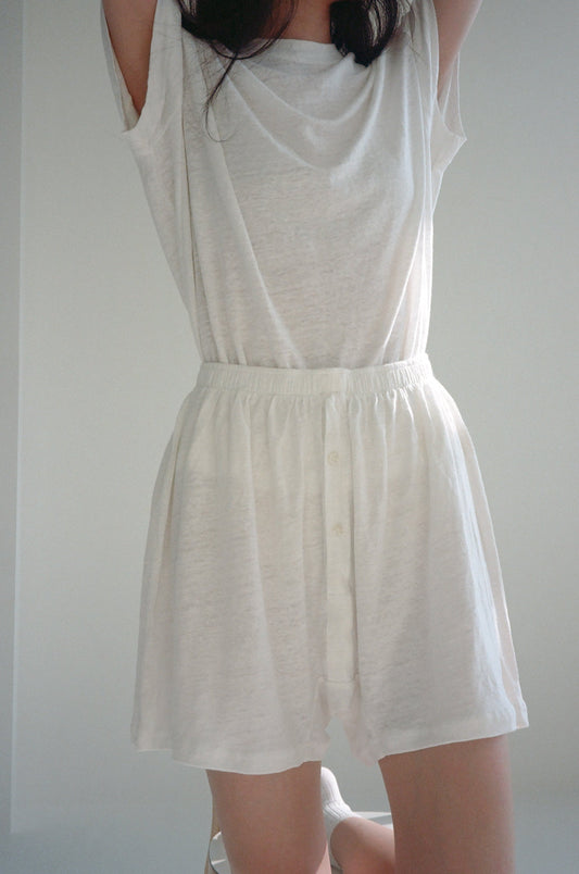 A person stands with arms raised against a plain light background, wearing high-waisted, button-front white Soft Shorts and a loose, short-sleeved, semi-sheer white top; shown from shoulders to knees.