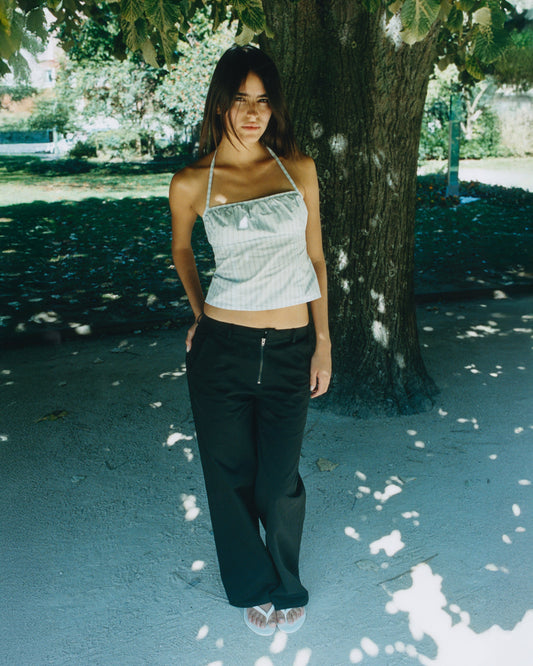 A woman stands under a leafy tree, wearing a light halter top and the Zip Pant - Black, crafted from organic cotton twill. Dappled sunlight patterns the ground and her clothes.