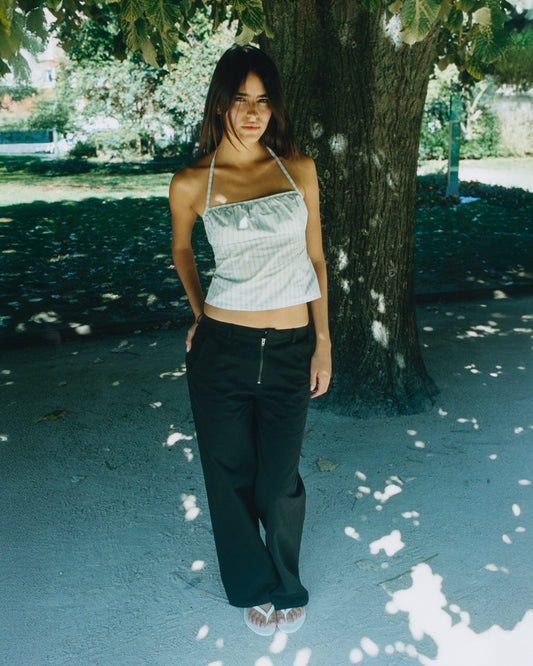 A woman stands outdoors under a large tree, wearing loose black Zip Pant, a light-colored halter top, and sandals as sunlight filters through the leaves, casting shadows on her outfit and the ground.