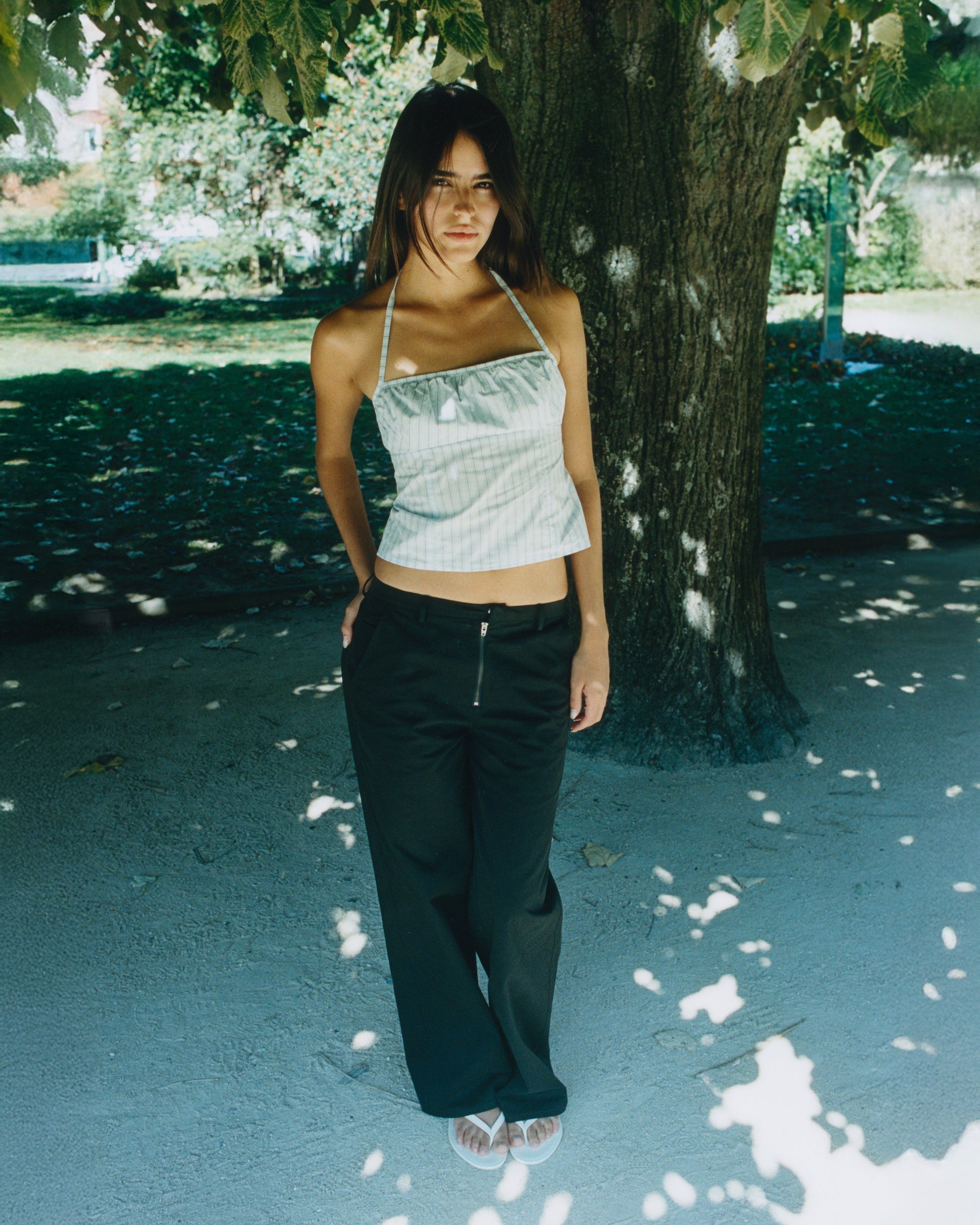 A woman stands outdoors under a large tree, wearing loose black Zip Pant, a light-colored halter top, and sandals as sunlight filters through the leaves, casting shadows on her outfit and the ground.