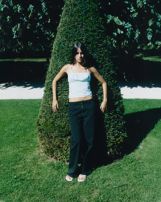 A woman stands in front of a cone-shaped bush in a sunny garden, wearing a relaxed fit light blue sleeveless top, white sandals, and the Zip Pant - Black. Shadows and lush greenery fill the background.