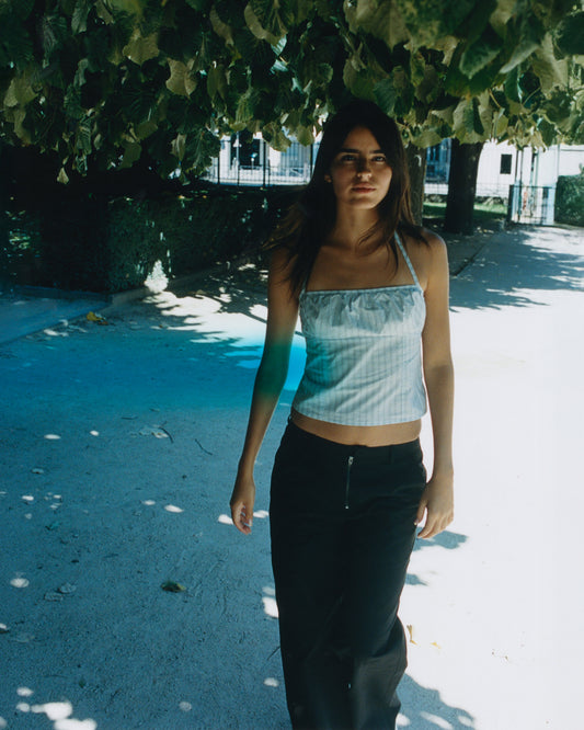A young woman with long brown hair stands under a leafy tree in dappled sunlight, wearing a light blue halter crop top and the Zip Pant - Black by Deiji Studios, set against an outdoor urban background.