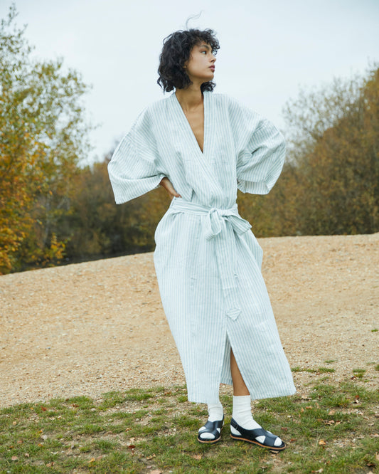Outdoors on a gravel path, a person wears the 02 Robe—a light blue, long, belted robe with wide sleeves—paired with white socks and black sandals. Autumn trees form the backdrop.
