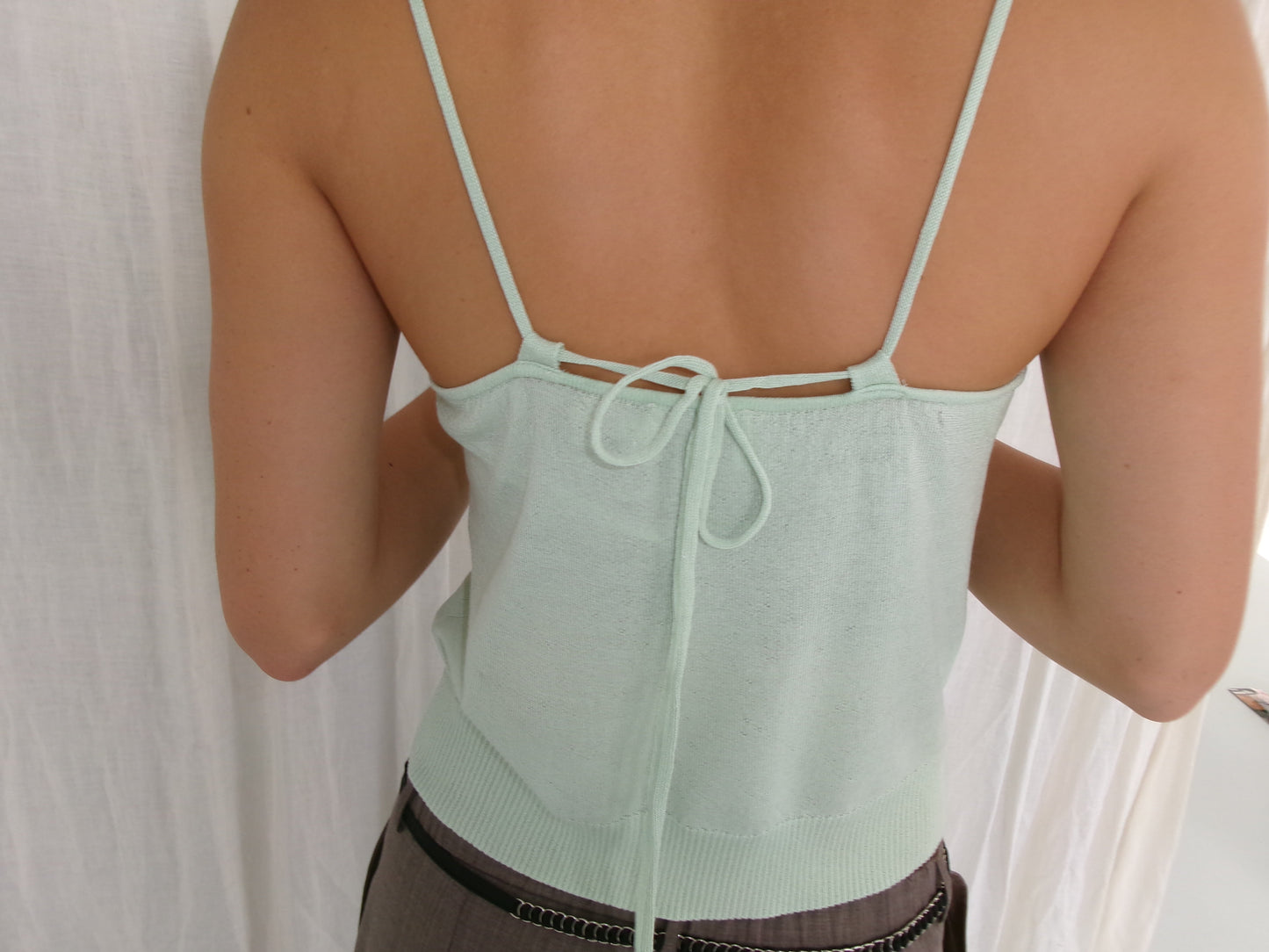 A person wears the Deiji Studios Drawcord Knit Top - Mint with thin straps and a bow tie at the back, featuring an adjustable ruched neckline. They stand against a white fabric backdrop, highlighting the open-back design.