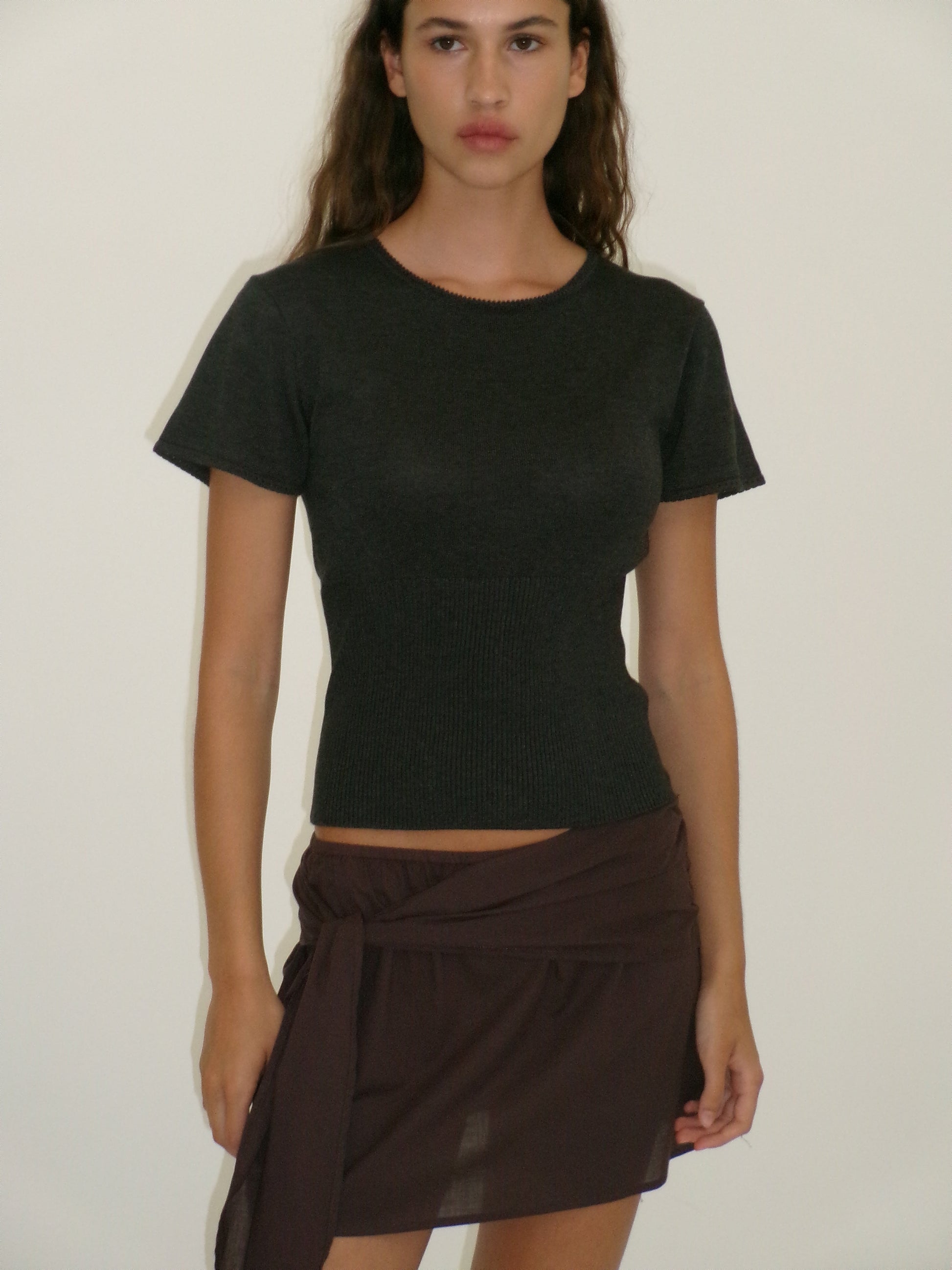 A woman with long brown hair wears a short-sleeved, fitted black top and the Belted Skirt - Wine from Deiji Studios, standing against a plain, light-colored background.