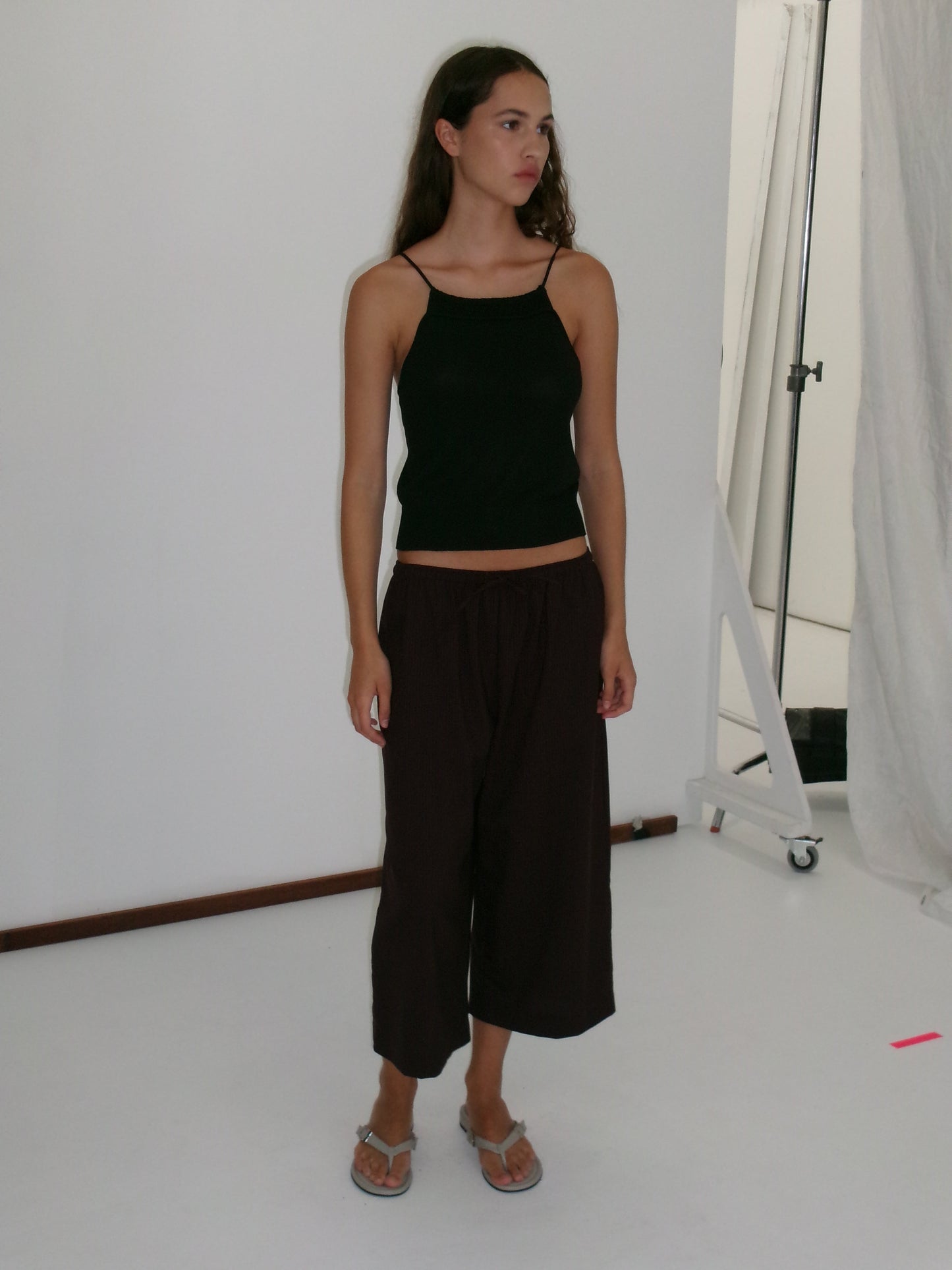 A young woman stands in a studio wearing a black sleeveless top and the Drawcord Culotte - Burgundy Brown Stripe from Deiji Studios with flip-flops. She has long hair, looks to the side neutrally, against a plain white background.