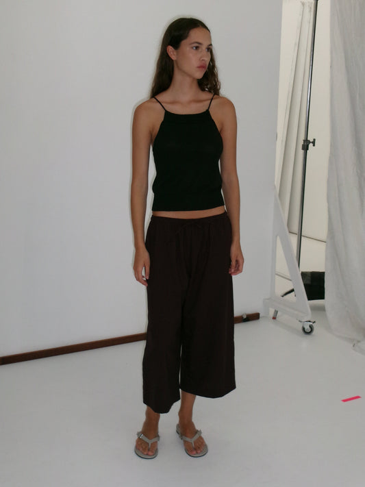 A young woman stands in a studio wearing a black sleeveless top and the Drawcord Culotte - Burgundy Brown Stripe from Deiji Studios with flip-flops. She has long hair, looks to the side neutrally, against a plain white background.