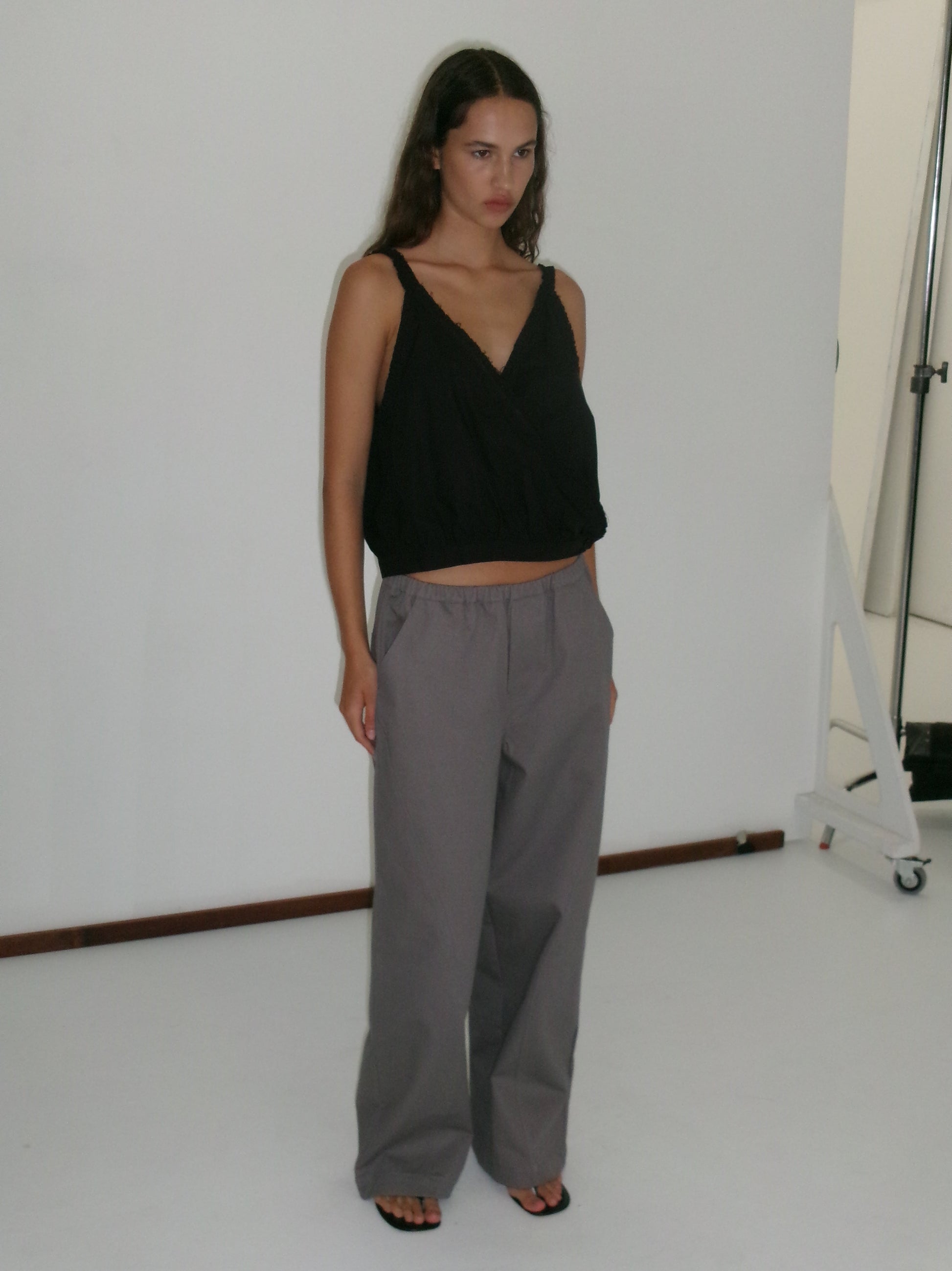 A woman with long hair and a neutral expression stands in a studio against a white background, wearing a sleeveless black V-neck top, Curve Pant - Lavender Smoke in a relaxed fit and mid-rise waist, and black sandals.
