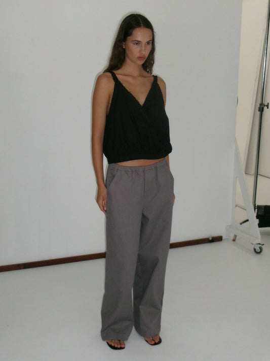 A woman with long hair and a neutral expression stands in a studio against a white background, wearing a sleeveless black V-neck top, Curve Pant - Lavender Smoke in a relaxed fit and mid-rise waist, and black sandals.