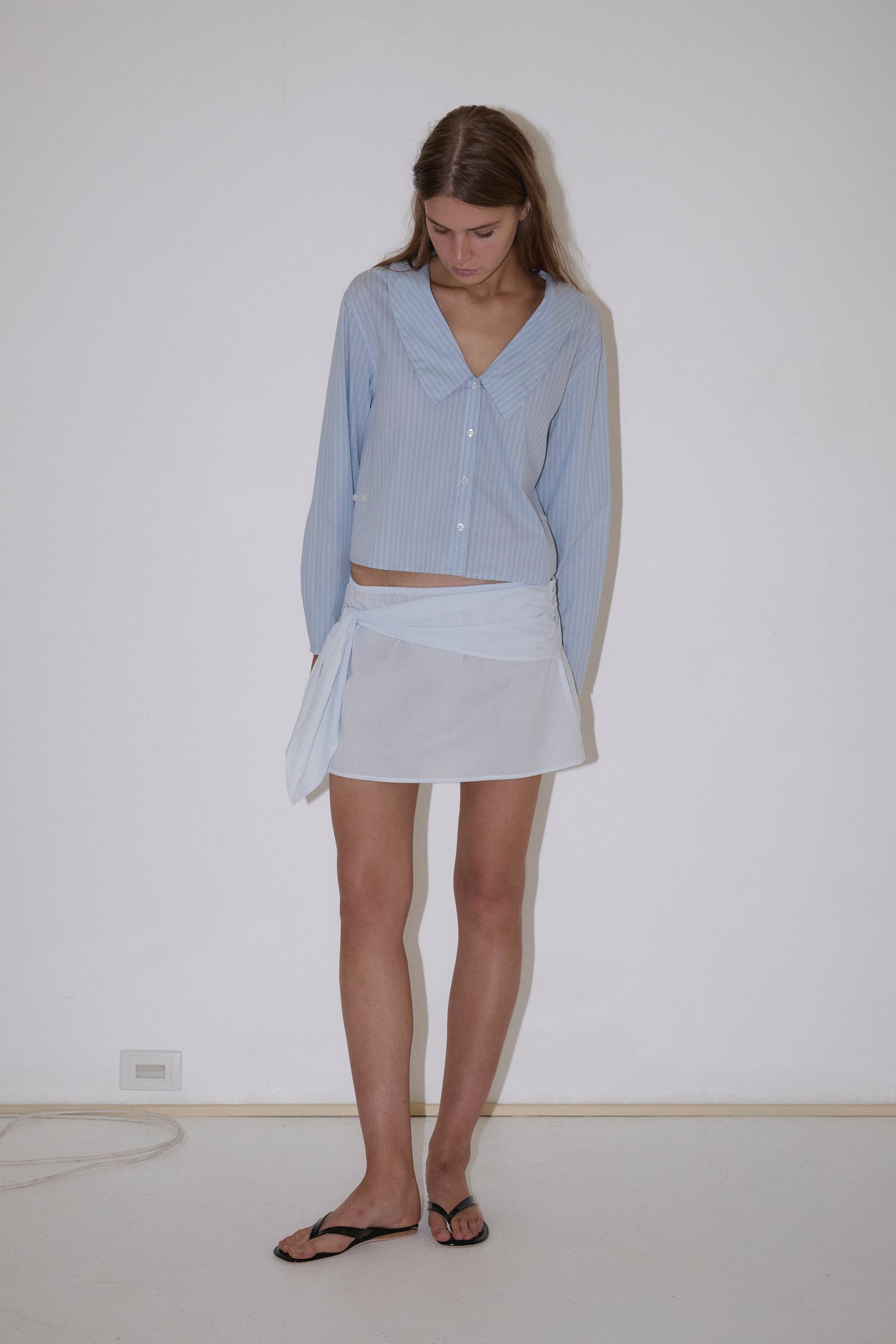 A woman stands indoors against a plain white wall, looking down. She wears a Deiji Studios Waist Button Shirt in Lake Blue Stripe, a white mini skirt with side tie, and black flip-flops.