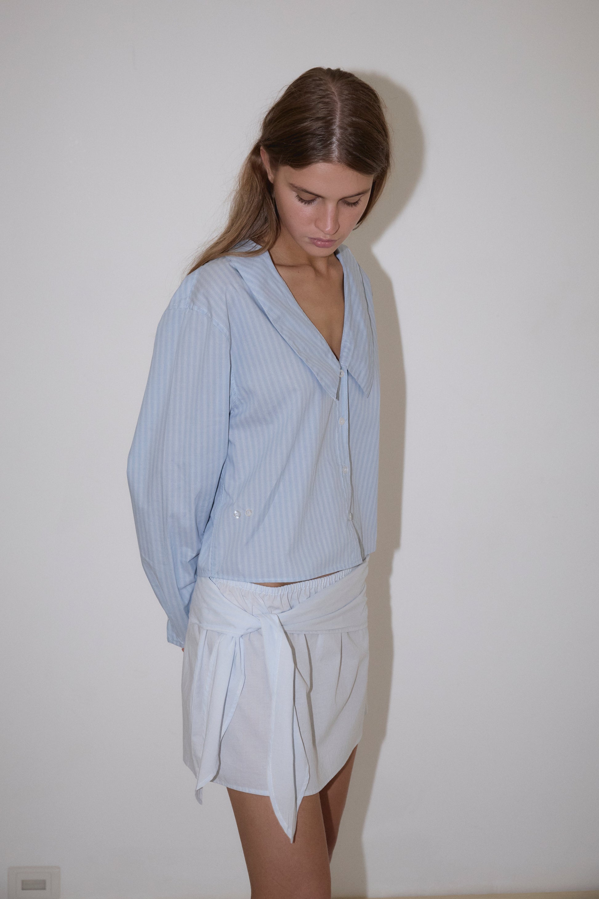 A woman with long brown hair stands against a white wall, looking down. She wears Deiji Studios' Waist Button Shirt in Lake Blue Stripe—a light blue, loose-fitting striped blouse—and a white wrap skirt, hands behind her back.