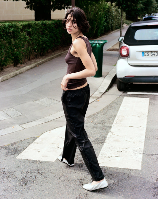 A person with short dark hair, wearing a dark tank top, Ruffle Pant, and white shoes stands on a crosswalk looking over their shoulder, with a hedge, green trash bin, and parked car in the background.