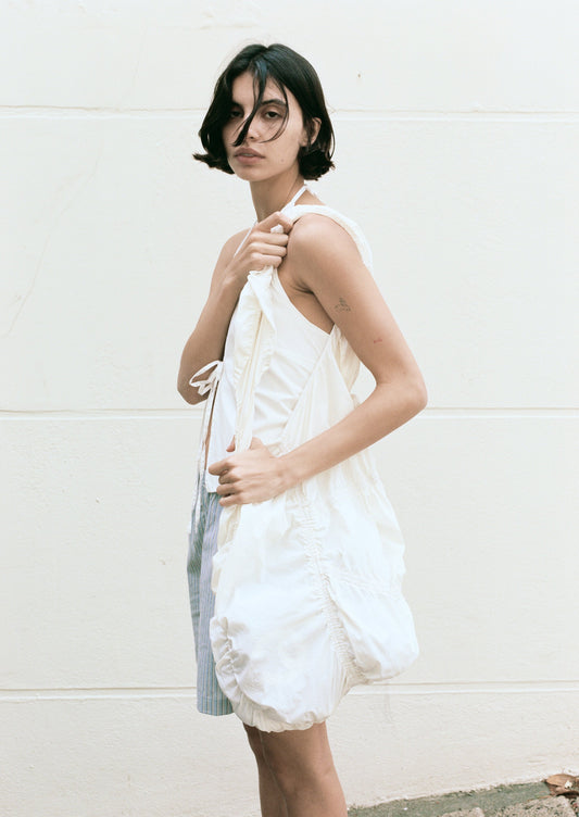 A person with short dark hair stands against a light wall, looking at the camera. They wear a sleeveless white dress with ruched details and carry the large, gathered Parachute Bag in matching white over their shoulder.