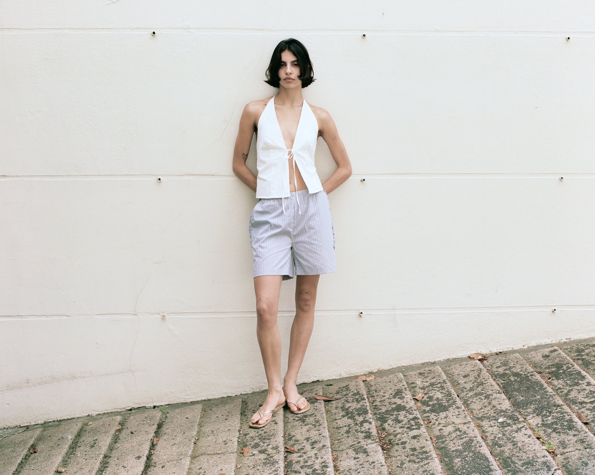 female model wears the open halter top top by Deiji Studios in white

top features open front with centre bust tie, halter neck tie and v neckline, bust seams for subtle shaping and shirred back

made from 100% GOTS certified organic cotton poplin

paired with a blue stripe cotton short