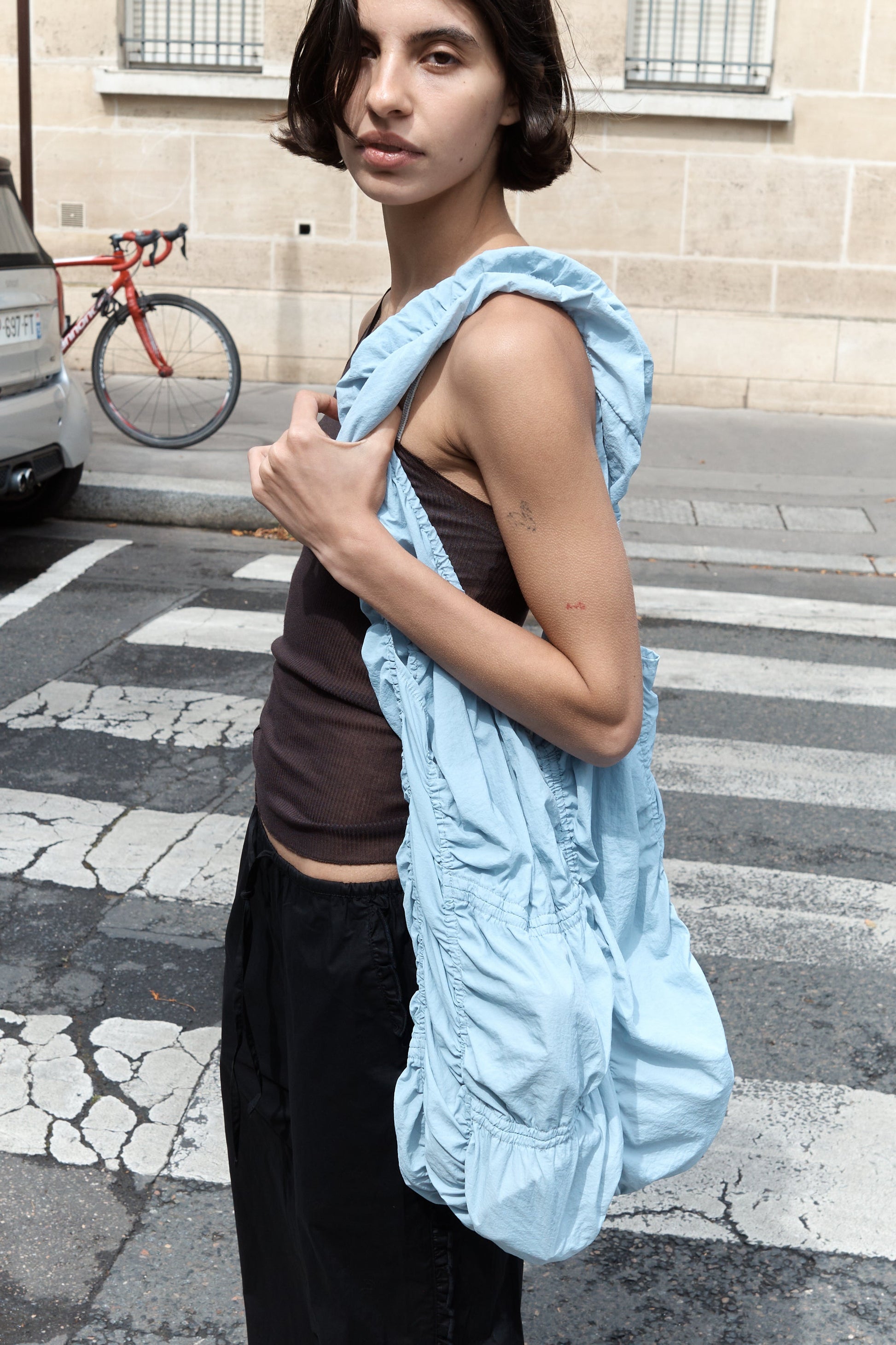 A person with short dark hair stands on a crosswalk, wearing a brown tank top, black pants, and carrying the Parachute Bag—a large ruched light blue shoulder bag. A red bicycle and beige building are visible in the background.