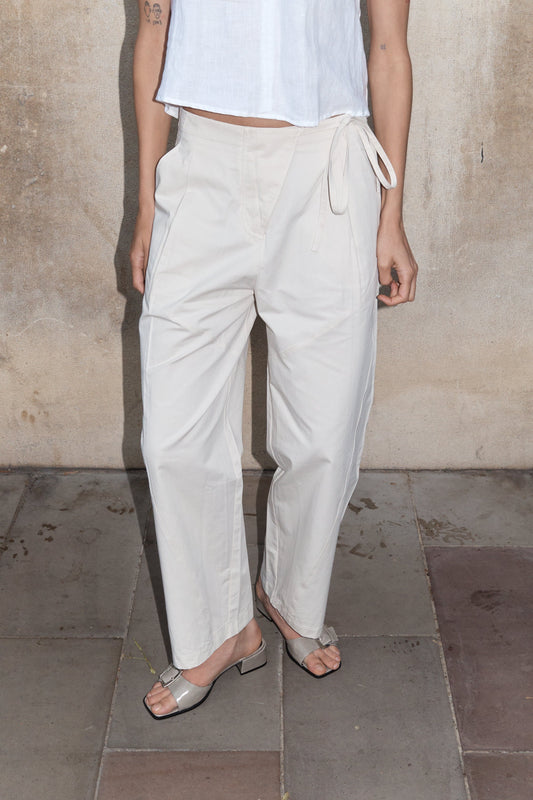 A person models the off-white Flag Tie Pant, styled high-waisted with a side tie, paired with a white sleeveless top and open-toe heeled sandals. They stand against a stone wall on a tiled floor; their face is not shown.