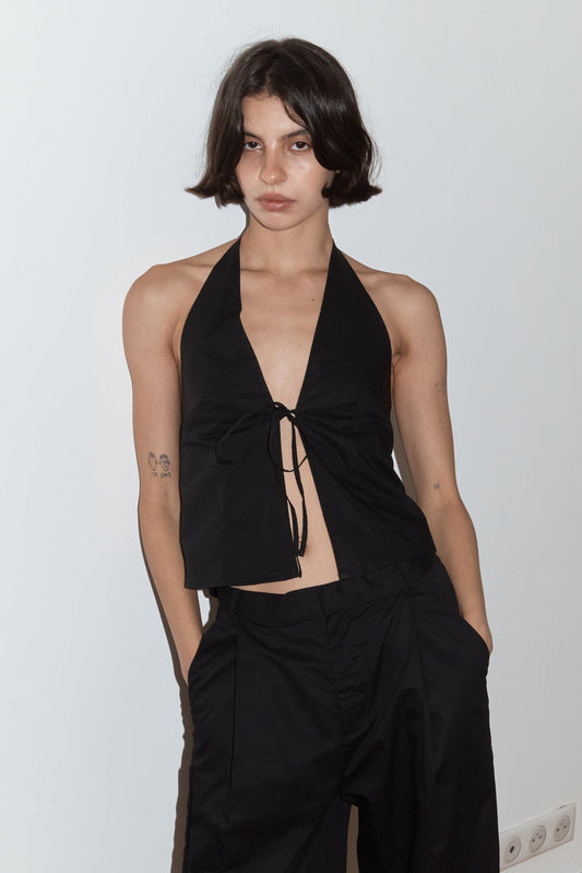 A model with short dark hair wears the Open Halter Top - Black from Deiji Studios, featuring an open halter design with tie-front detail, paired with black wide-leg trousers, posed against a white wall.