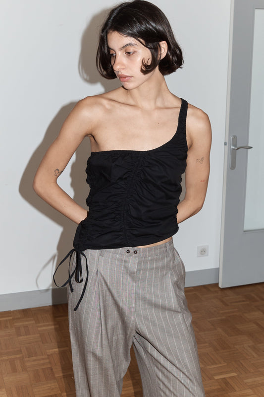 A person with short dark hair wears the Scrunchie Top - Black from Deiji Studios—an asymmetrical organic cotton one-shoulder top—paired with loose gray pinstriped pants, standing indoors by a white wall and door, looking slightly down and to the side.