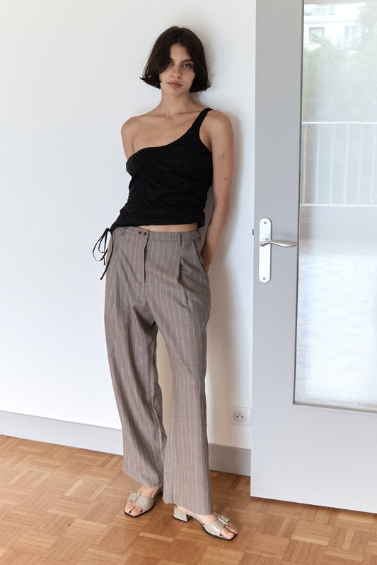 A woman with short dark hair stands indoors on a wooden floor by a white door, wearing a black asymmetrical top and beige Twin Button Pant.