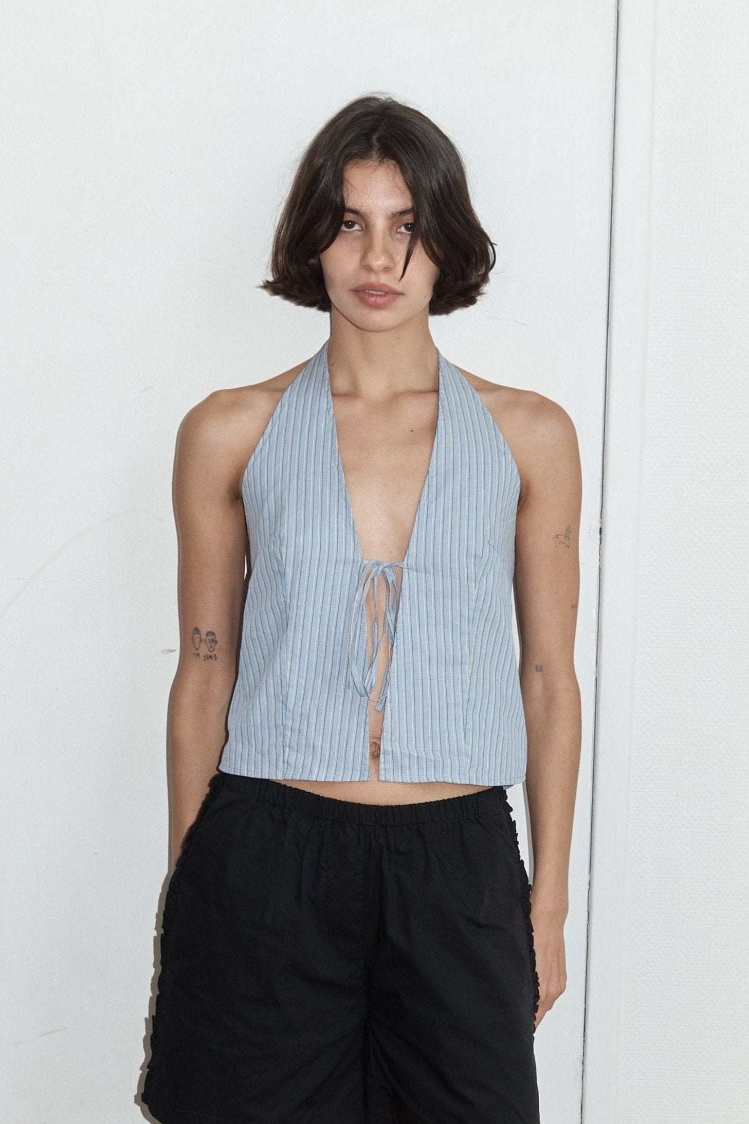 A person with short dark hair models the Open Halter Top, a sleeveless light blue V-neck with a front tie, paired with loose black shorts, standing against a plain white background.