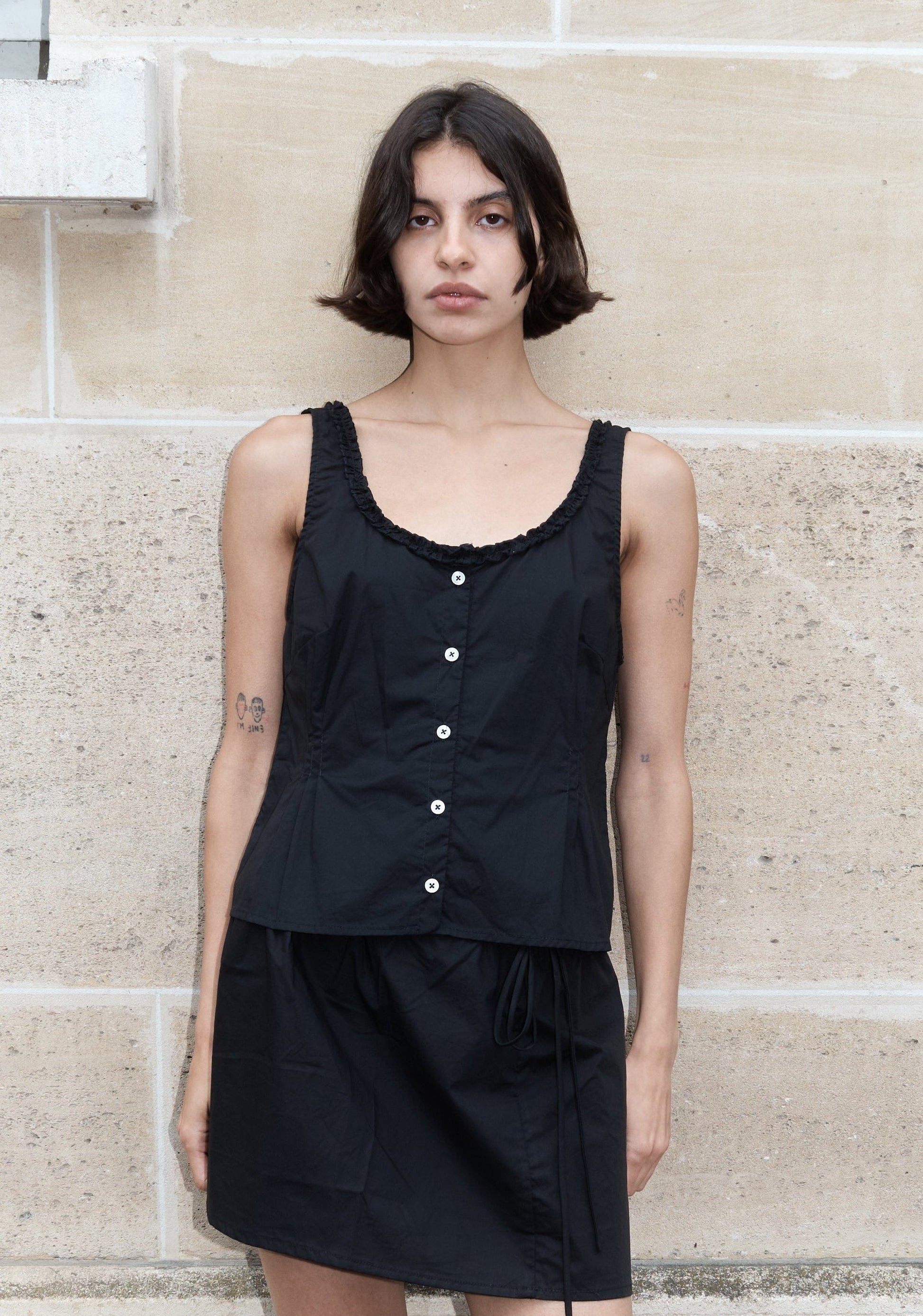 A woman with short dark hair and visible arm tattoos stands against a beige stone wall, wearing the Ruffle top—a sleeveless black top with white buttons—paired with a matching black skirt. She has a neutral expression.