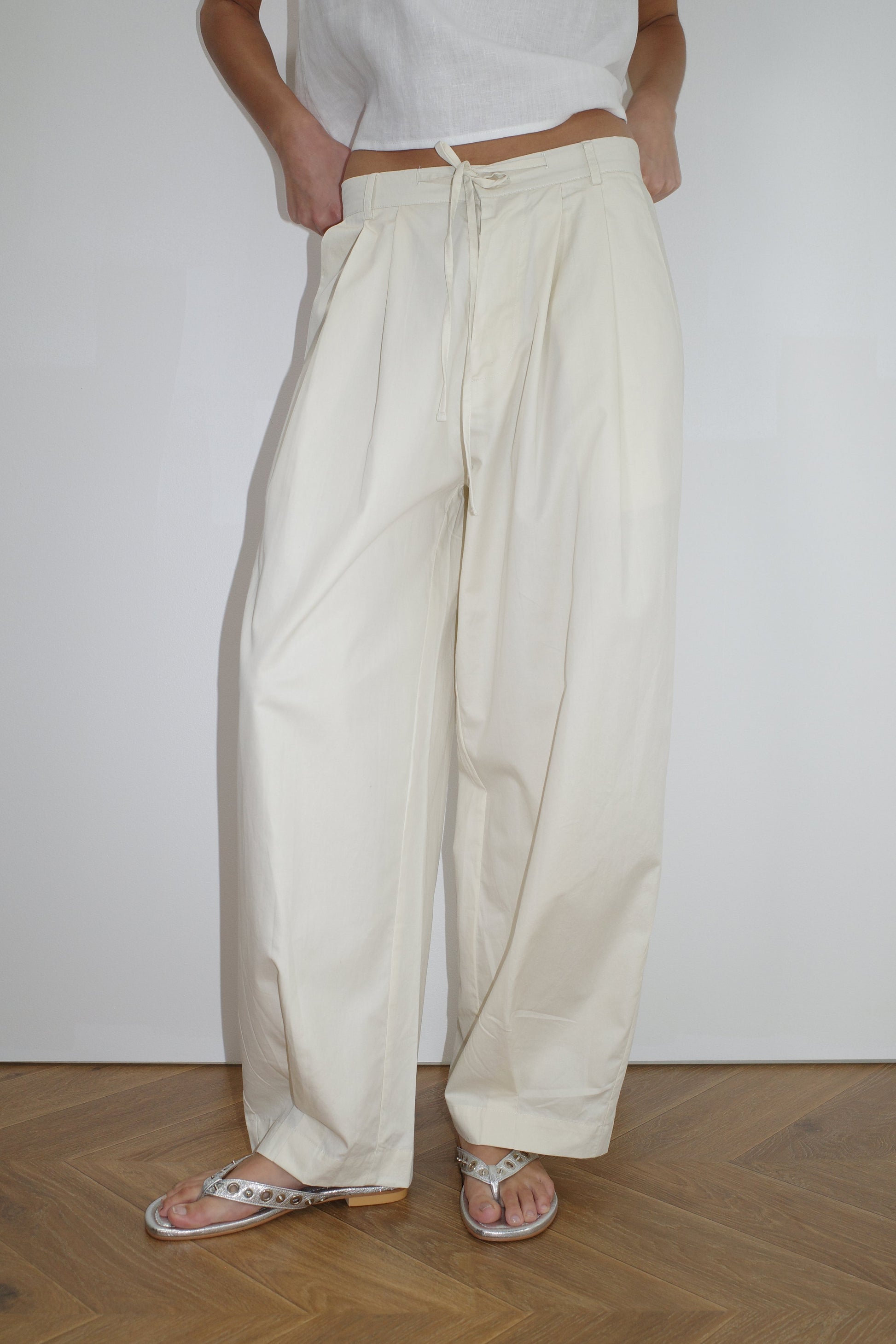 A person wears the Turn Pant - Off White by Deiji Studios—high-waisted, relaxed fit with an adjustable drawcord—paired with a white top and sandals, standing on a wooden floor in front of a plain white wall.