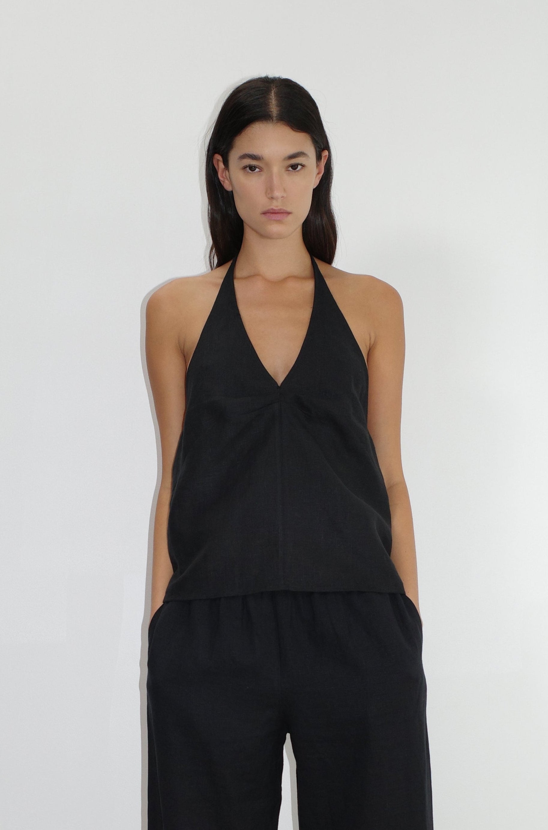 A person with long dark hair stands against a white background, wearing the Linen Halter Top—a sleeveless black V-neck halter—with matching loose black pants, looking at the camera with hands in pockets.
