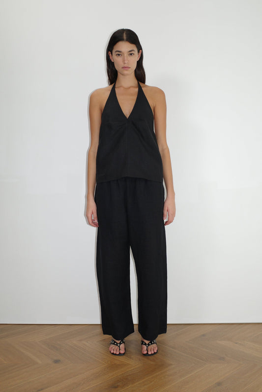 A woman stands against a plain white wall wearing loose-fitting black Linen Ease Trousers, a sleeveless black halter top, and black sandals on a wooden floor, facing forward with her arms relaxed at her sides.
