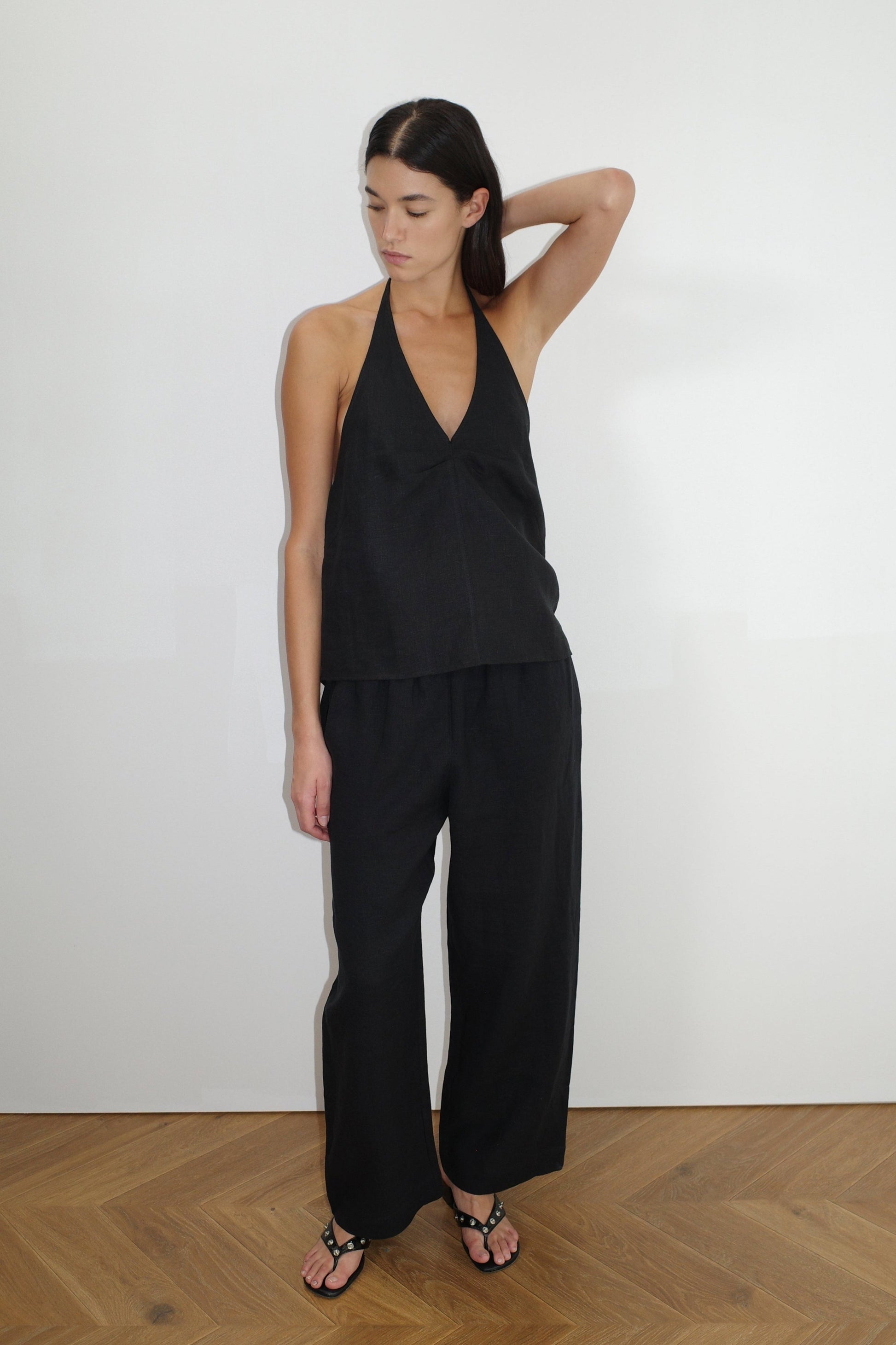 A woman with straight dark hair stands indoors against a white wall, wearing the Linen Ease Trouser - Black with a sleeveless halter top and black sandals. She looks down, one arm raised and bent behind her head.
