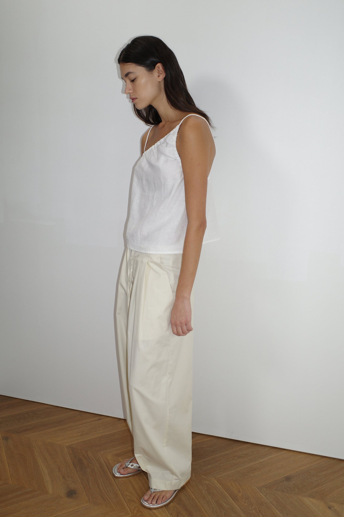 female model wears the turn pant in off white, a relaxed tailored pant features soft pleating at the front, buttoned waistband and adjustable inner drawcord. designed with hidden side seam pockets and pockets at the back, made from 100% GOTS certified organic cotton paired with a white top
