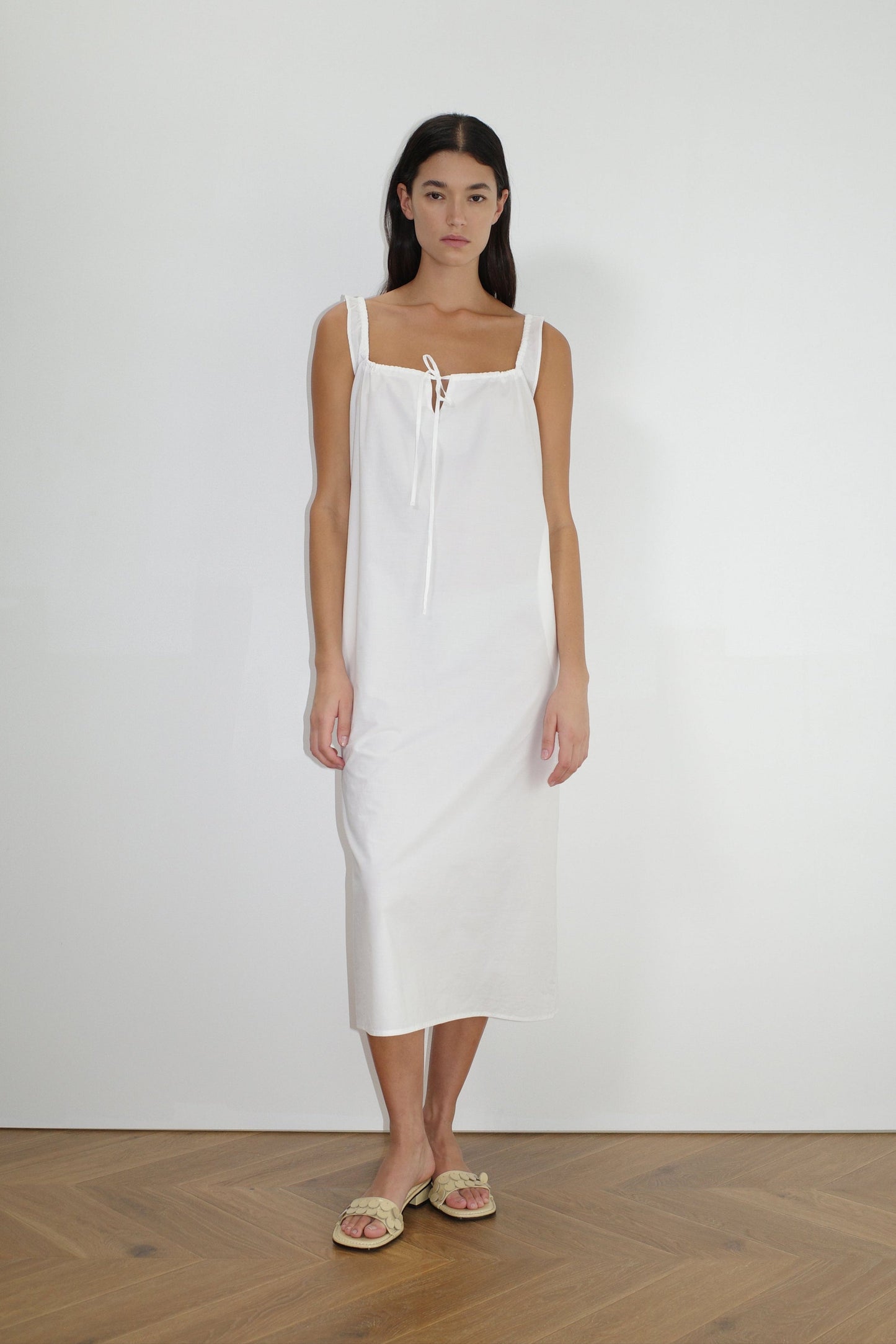 A woman with long dark hair and a neutral expression stands indoors against a plain white wall, wearing The Paper Dress - White by Deiji Studios, a sleeveless square-neck maxi dress with tie details, paired with light-colored sandals.